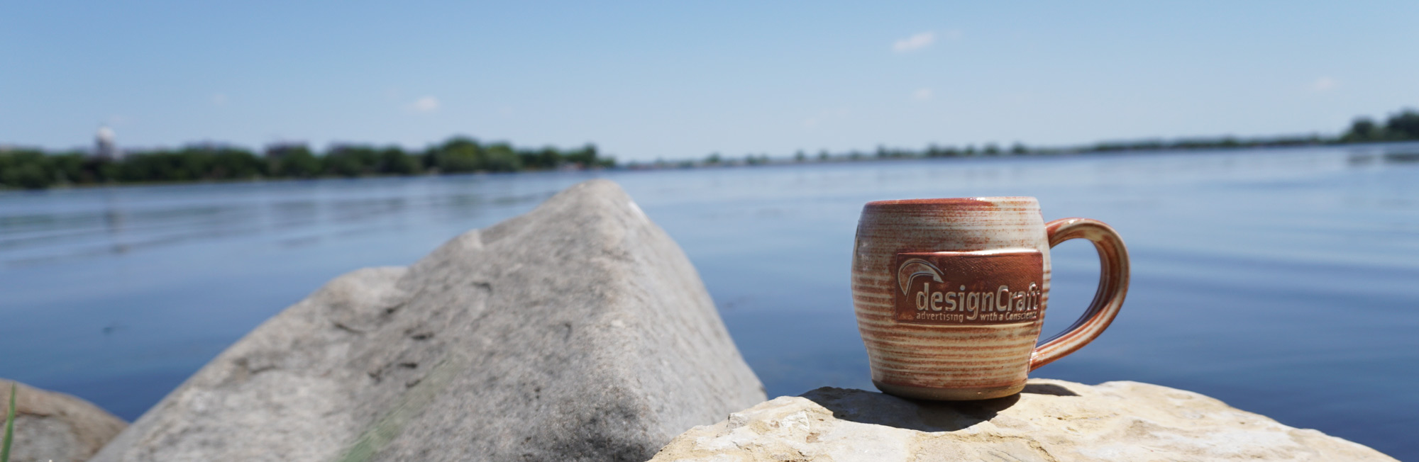 Handmade pottery mug with designCraft logo on it sitting on rock by lake